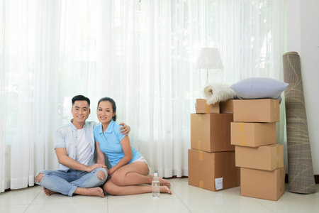 Young Asian couple embracing on floor of new spacious flat with boxes near smiling at camera