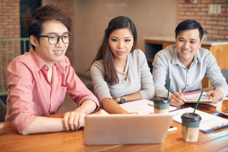 Group of modern Asian men and woman sitting at table in cafe having coffee break and watching laptopの写真素材