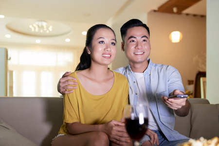 Young Asian couple sitting on sofa with wineglasses and watching TV at home