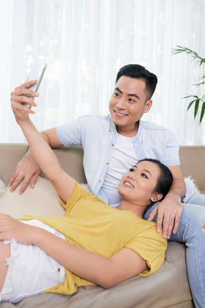 Cheerful young Asian couple sitting on couch at home and taking selfie with phone togetherの写真素材