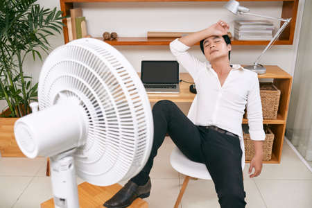 Young businessman suffers from heat while working in the office and tries to cool off by the fanの写真素材
