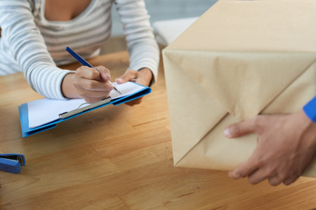 Crop shot of woman in post service signing paper on clipboard and receiving parcel at table