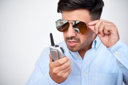 Confident young man in sunglasses talking on radio set while standing isolated on white