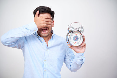 Portrait of man in shirt holding alarm clock and screaming while covering his face with hands...