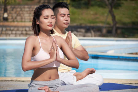 Young Asian couple sitting in lotus position with eyes closed and meditating near the swimming pool outsideの写真素材