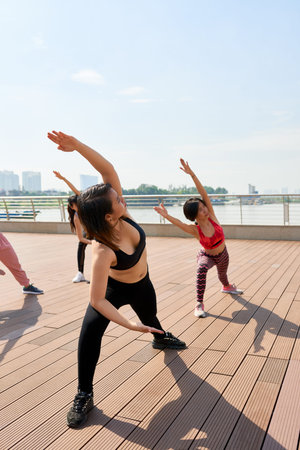 Sportive ethnic women in sportswear working out on city seafront in bright sunlight doing exercise...