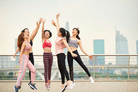 Group of laughing Asian woman in sportswear jumping and giving high five against cityscapeの写真素材