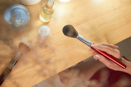 Close-up of young woman sitting at the table with tubes with cream and holding make-up brush, she is...