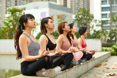 Young sportive Asian women sitting in row on pond fence in park and meditating peacefullyの写真素材