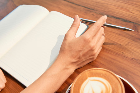 Close-up of woman sitting at wooden table and writing plans in her notebook while drinking coffee