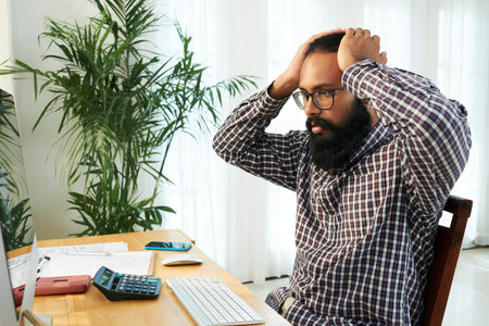 Shocked young programmer keeping his hands on head while looking at data on computer screen