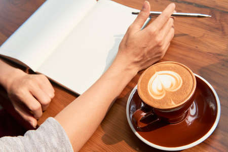 Close-up of female hands holding pen and sitting at the table in front of open blank notepad with cup of cappuccinoの写真素材