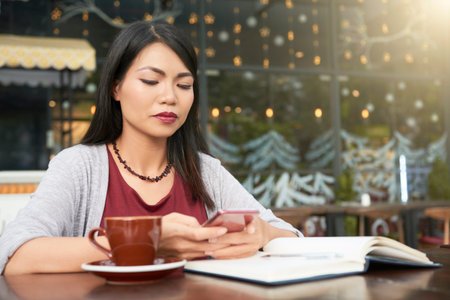 Young attractive woman sitting at the table with notepad and cup of coffee and using her smartphone...