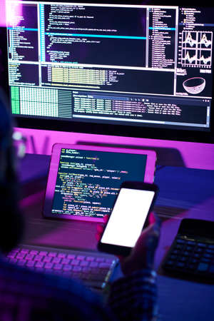 Modern programmer with smartphone sitting by workplace in front of tablet and computer monitor with coded dataの写真素材