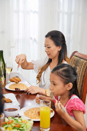 Beautiful Asian woman putting food on plate while sitting with family at table at homeの写真素材