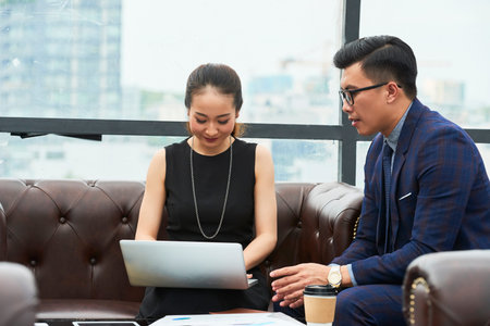 Smiling pretty Vietnamese businesswoman working on laptop when listening to ideas of her colleagueの写真素材