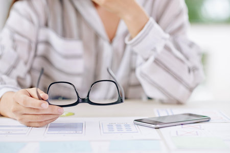Young tired analyst holding eyeglasses in hand over paper schemes and smartphone while sitting by...