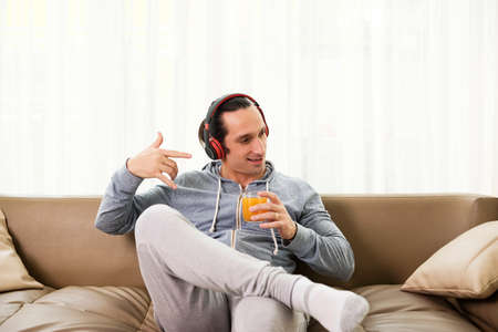Young man sitting on sofa with headphones listening to music and drinking fresh fruit juice at homeの写真素材