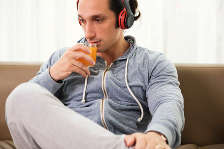 Handsome man holding glass with orange juice and drinking it while listening to music in headphones...