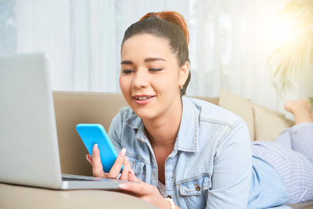 Smiling young woman lying on the sofa in front of laptop computer and using her smartphone for...