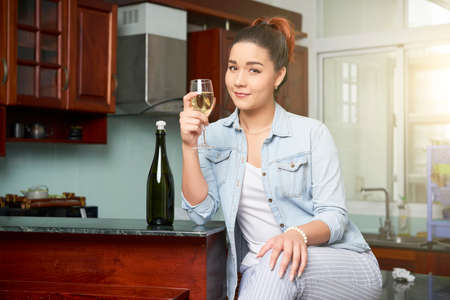 Portrait of beautiful woman holding a glass with white wine while sitting in the kitchen at homeの写真素材