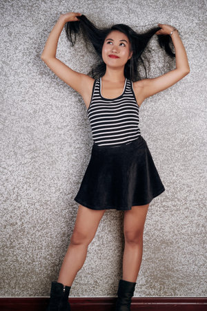 Young Asian lady in stylish skirt holding her dark long hair against the wall