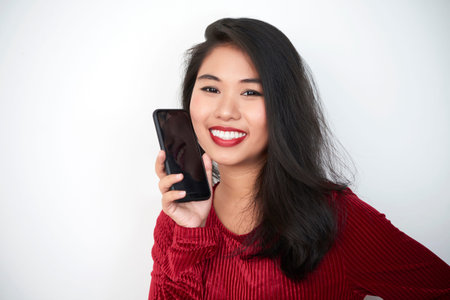 Portrait of Asian pretty brunette in elegant blouse holding mobile phone and smiling at camera...