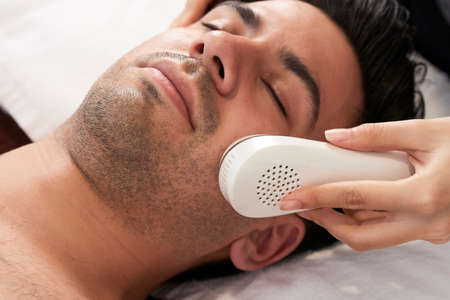 Close-up of male face with eyes closed and cosmetologist working with ultrasonic equipment on his faceの写真素材