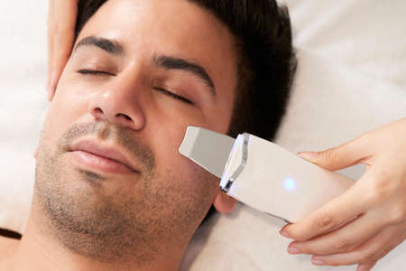 Close-up of cosmetologist doing ultrasound face scrubber on male face at cosmetology clinicの写真素材