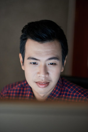 Positive Vietnamese man reading information on computer screen when working at night