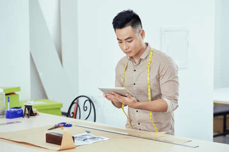 Young Vietnamese tailor standing at workplace and watching tutorial on tablet computerの写真素材