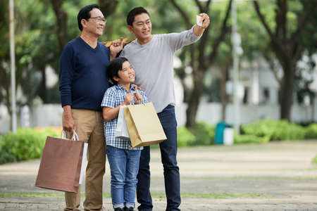 Mature Vietnamese man taking selfie with his father and son after shoppingの写真素材