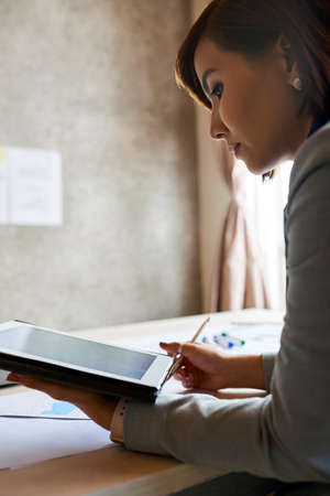 Successful young female entrepreneur reading information on digital tablet and taking notes when working in officeの写真素材