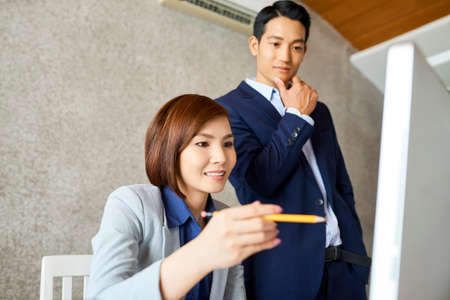 Smiling Asian coworkers looking at computer screen and discussing progressの写真素材
