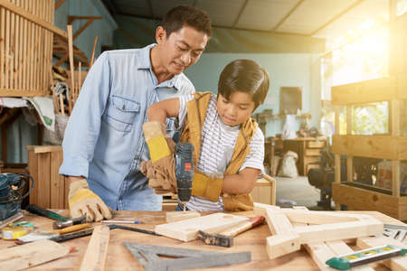 Preteen Vietnamese boy drilling wooden plancks in workshop under control of his fatherの写真素材