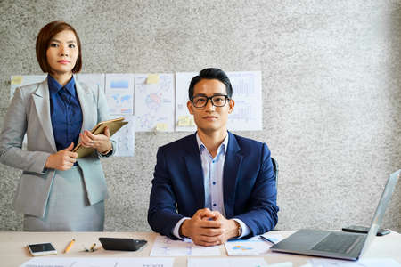 Handsome young Vietnamese businessman and his pretty assistant looking at cameraの写真素材