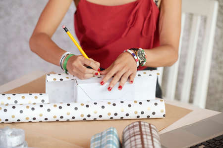 Close-up of woman wrapping present in gift paper and signing it with pencil at the tableの写真素材