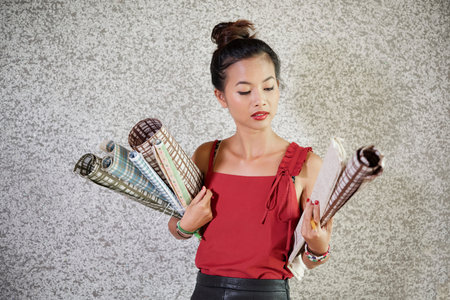 Asian young woman standing and holding colourful wrapping paper rolls for gifts against grey...