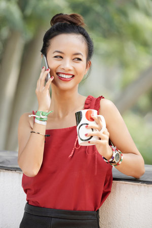 Young happy Asian woman standing and drinking coffee during conversation on mobile phone outdoors
