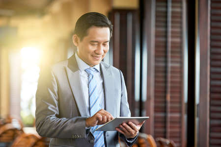 Asian mature business manager in formal wear standing and smiling while typing on tablet pc in cafeの写真素材