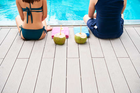 Rear view of young couple sitting and resting near the poolside outdoors with coconut water drinksの写真素材
