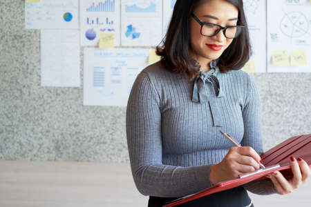 Asian pretty financial advisor in eyeglasses making notes in her red folder while visiting financial presentation at officeの写真素材