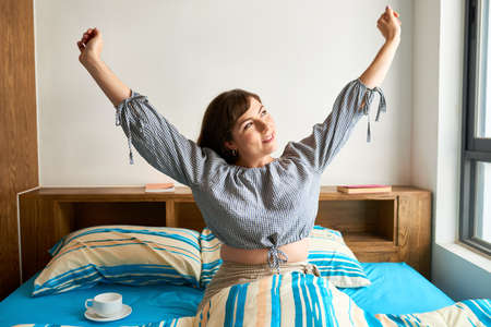 Happy pretty woman stretching in her bed and enjoying new morningの写真素材