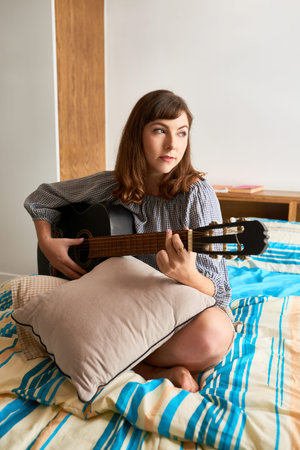 Lovely young woman sitting on bed with guitar and singing a song