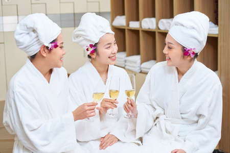 Group of Asian young women in bathrobes drinking champagne and talking while sitting in dressing roomの写真素材