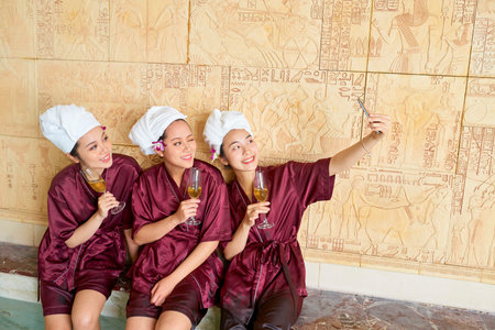 Group of young Asian girls sitting in gowns with glasses of champagne and making selfie on mobile...