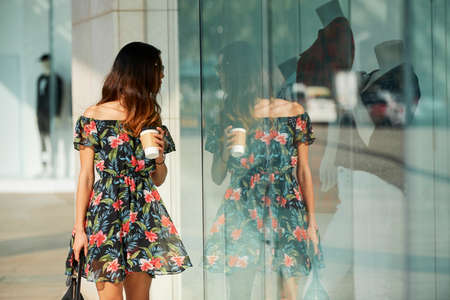 Young female in elegant floral dress holding cup of hot drink and looking at clothing behind shop window while standing on city street on sunny dayの写真素材