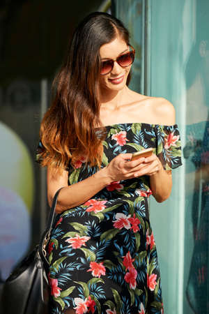 beautiful Asian female in floral dress smiling and using modern smartphone while standing near shop window on city street on sunny dayの写真素材