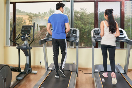 Rear view of young couple running on treadmills together in front of big window in fitness club