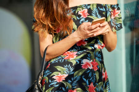 Anonymous female in floral dress browsing smartphone while standing on city street on sunny dayの写真素材